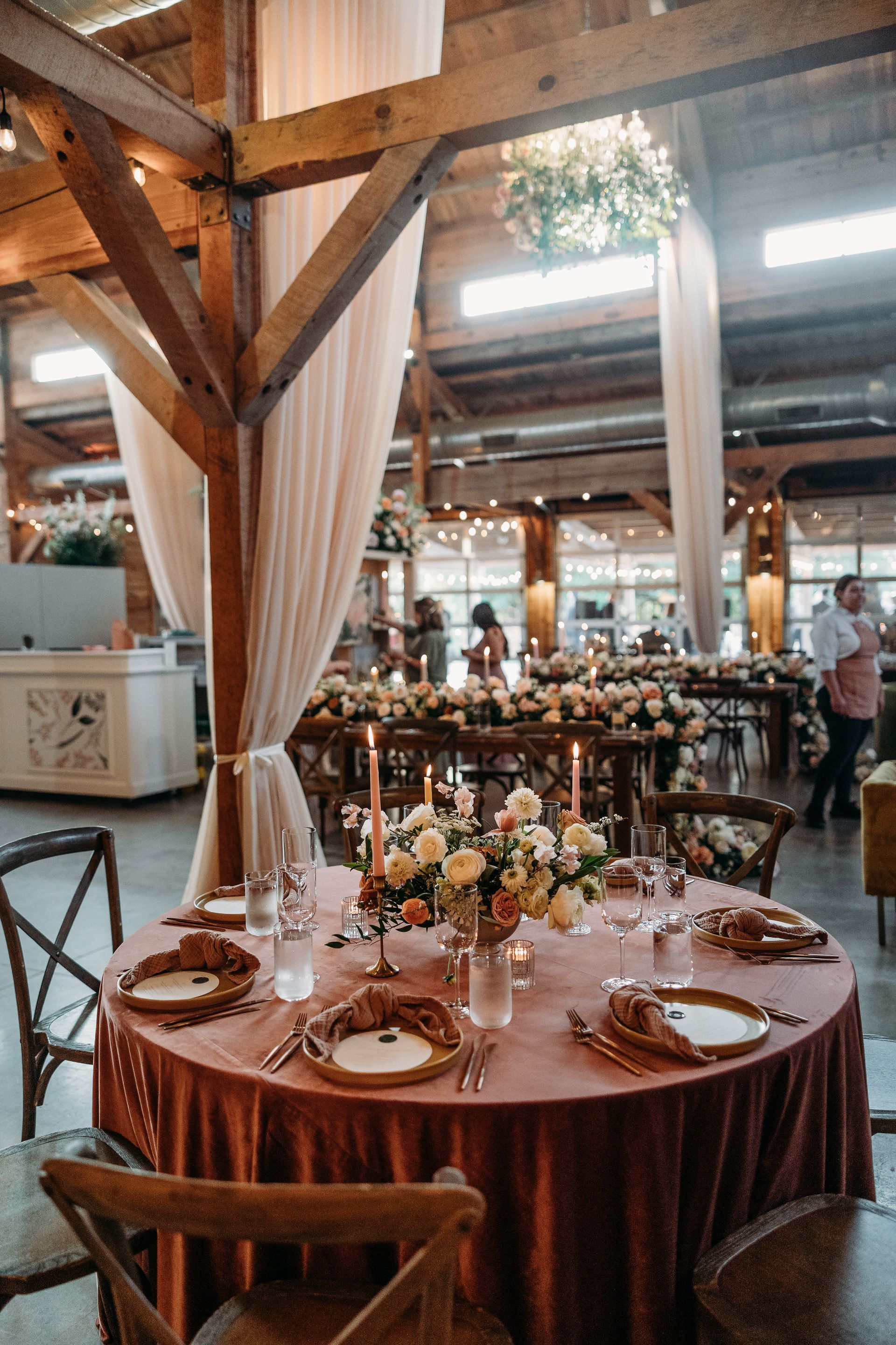 Rustic banquet hall with a set round table, floral centerpiece, and guests in the background
