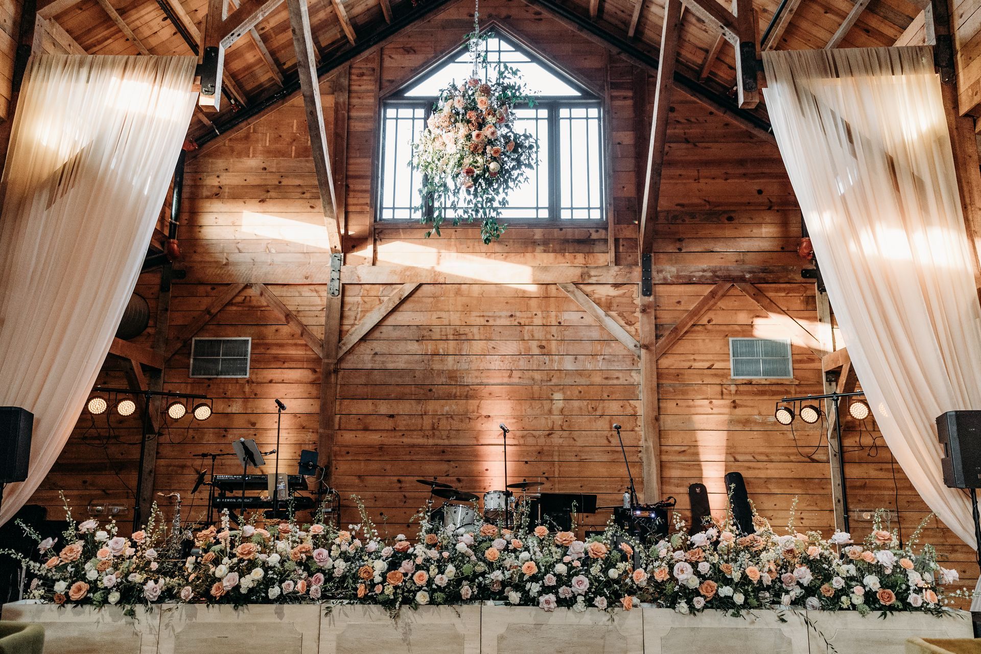 Rustic barn wedding ceremony setup with white drapes, floral arrangements, and hanging greenery under a wood beam ceiling