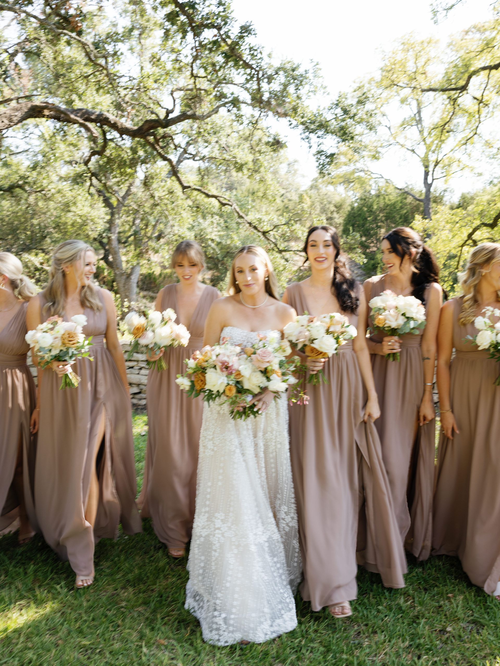 Bridal party in taupe dresses holding bouquets outdoors under a leafy tree