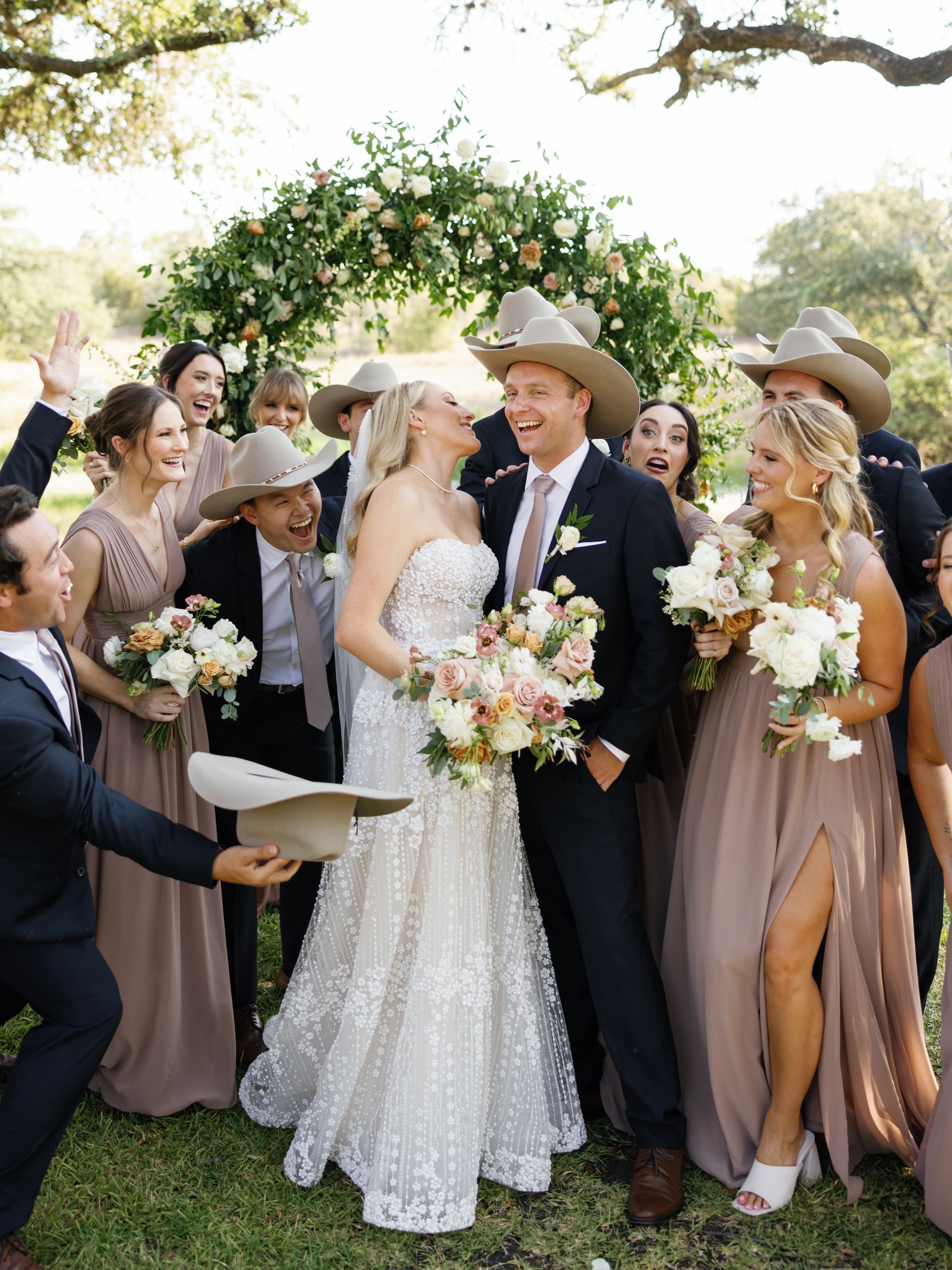 Wedding party gathered around bride and groom under a flower arch outdoors.