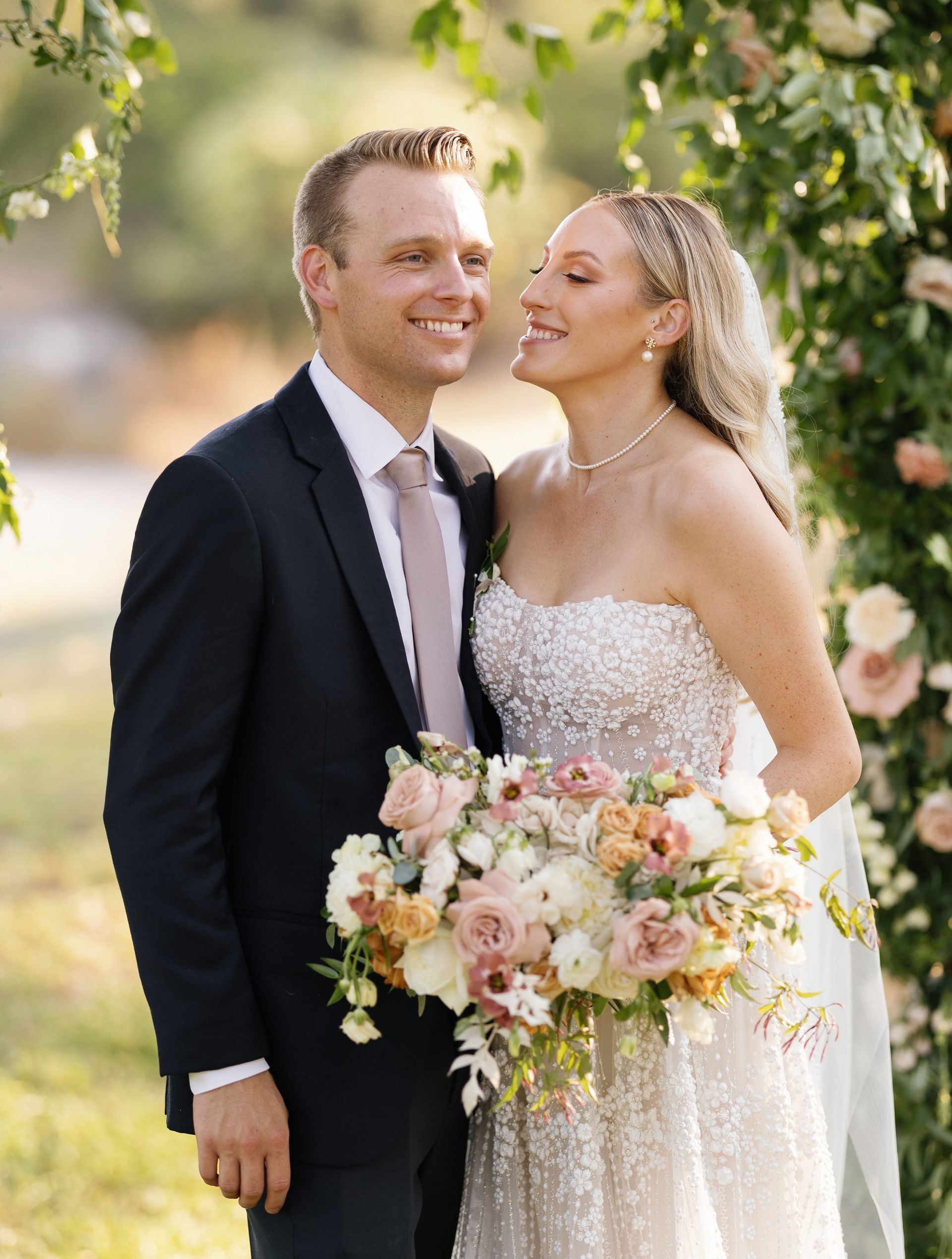 Bride and groom smiling outdoors, bride holding pastel bouquet in a wedding dress and groom in a dark suit