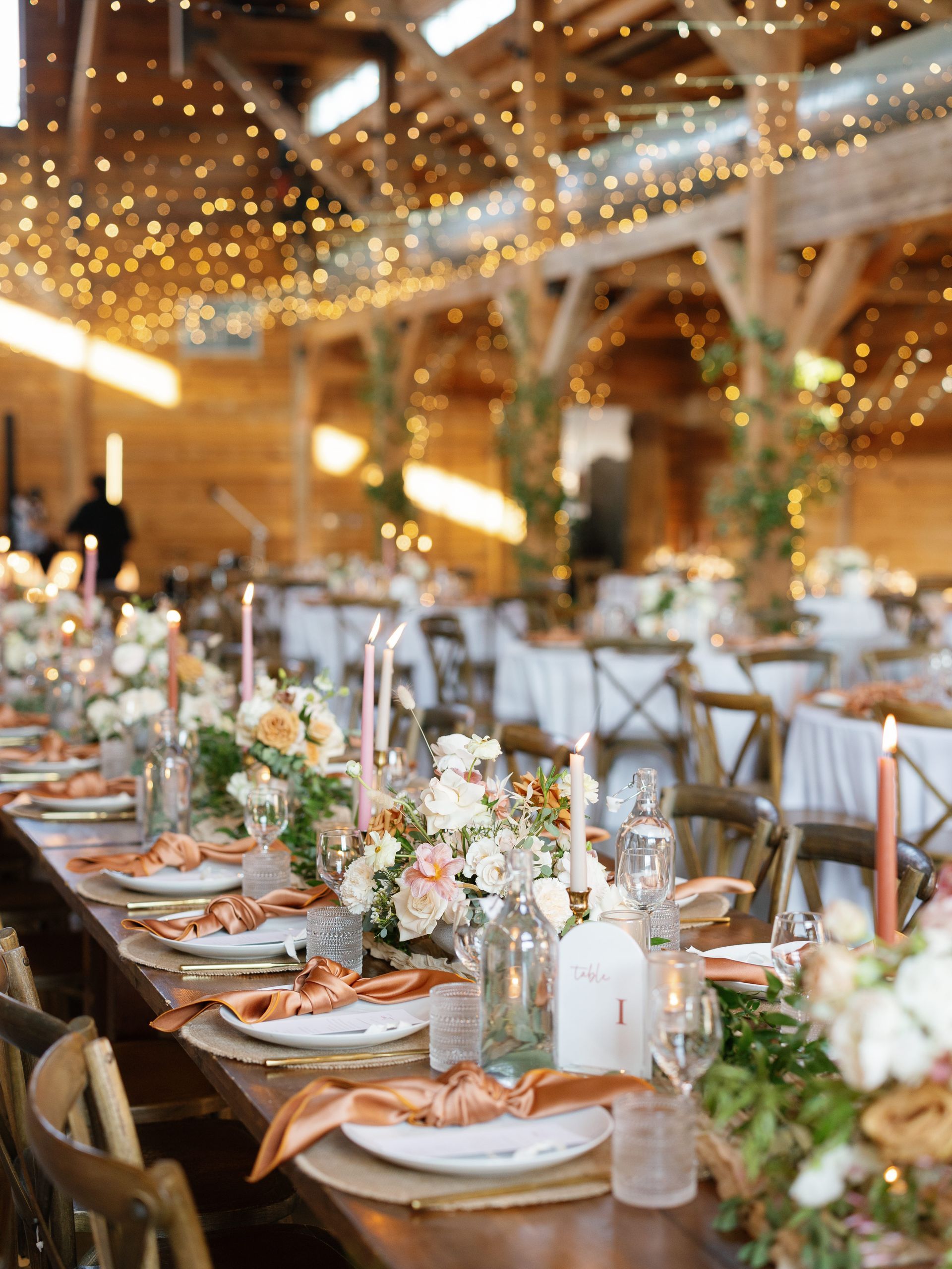 Elegant banquet table with white chairs, floral centerpieces, candles, and string lights in a rustic venue