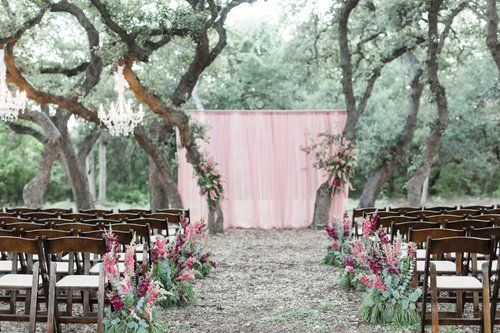 Outdoor wedding aisle with pink floral decorations, wooden chairs, and pink drapery backdrop under trees