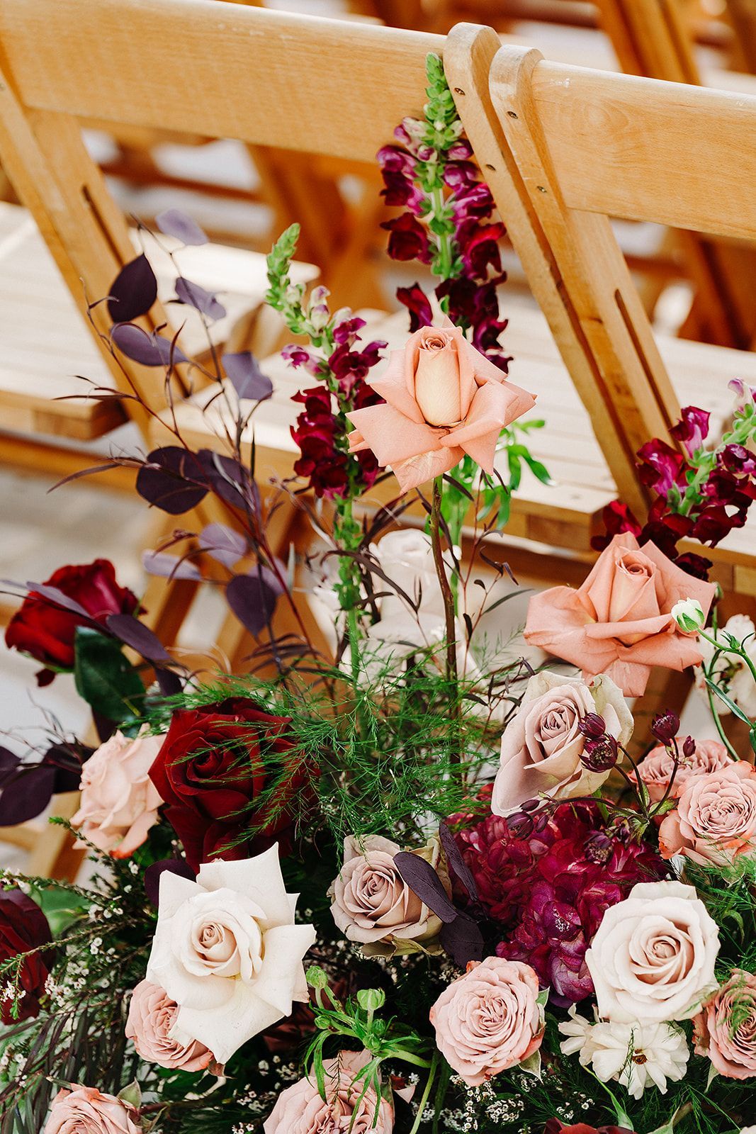 Floral arrangement with pink, cream, and burgundy flowers beside wooden chairs at a wedding-like setting