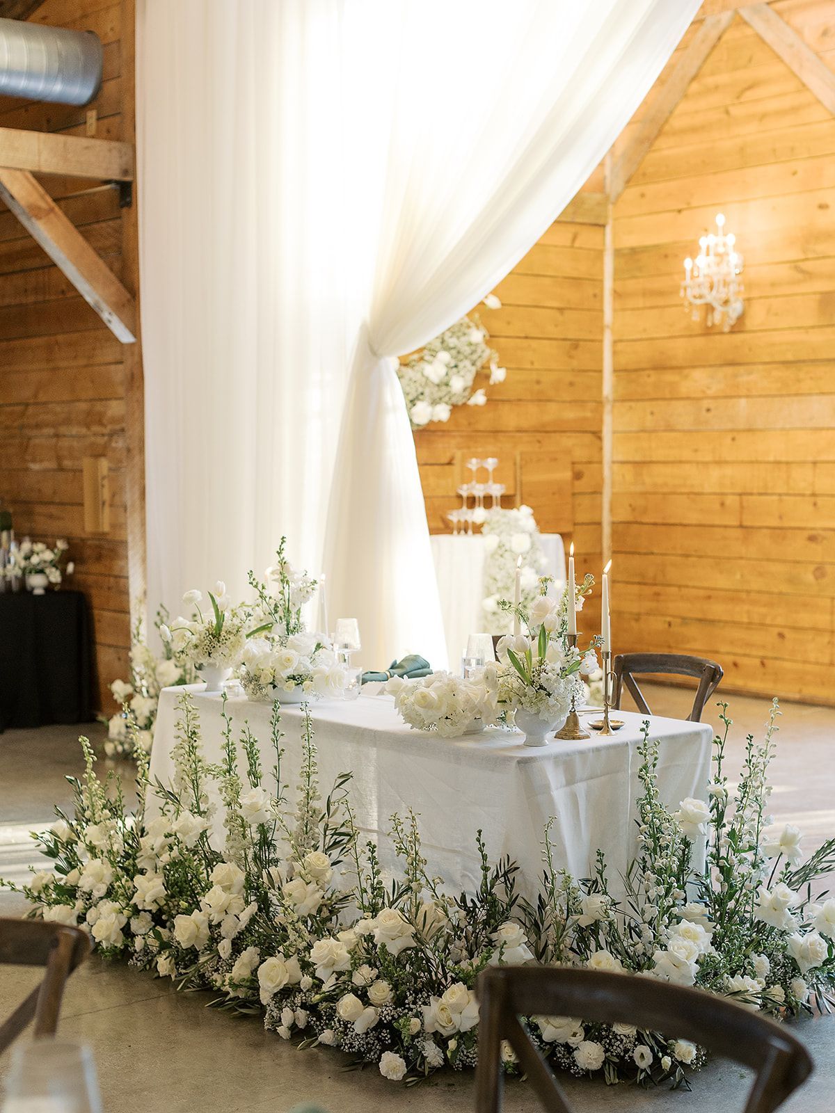 White floral wedding reception table in a wooden hall with draped curtains and candles