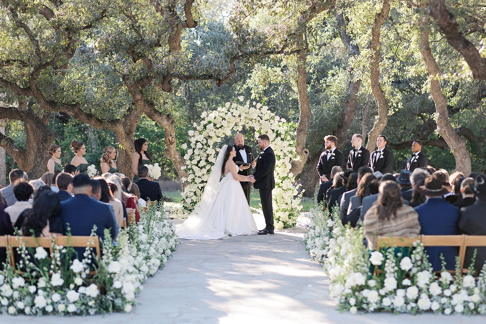 Outdoor wedding ceremony under leafy trees, bride and groom at the altar, guests seated on either side.