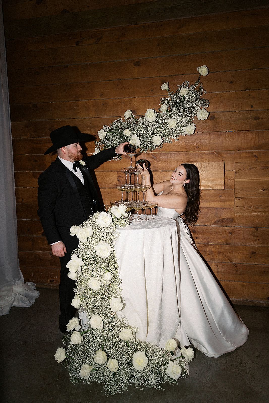 Couple cutting a wedding cake on a rustic wooden backdrop, with white floral garland and candles