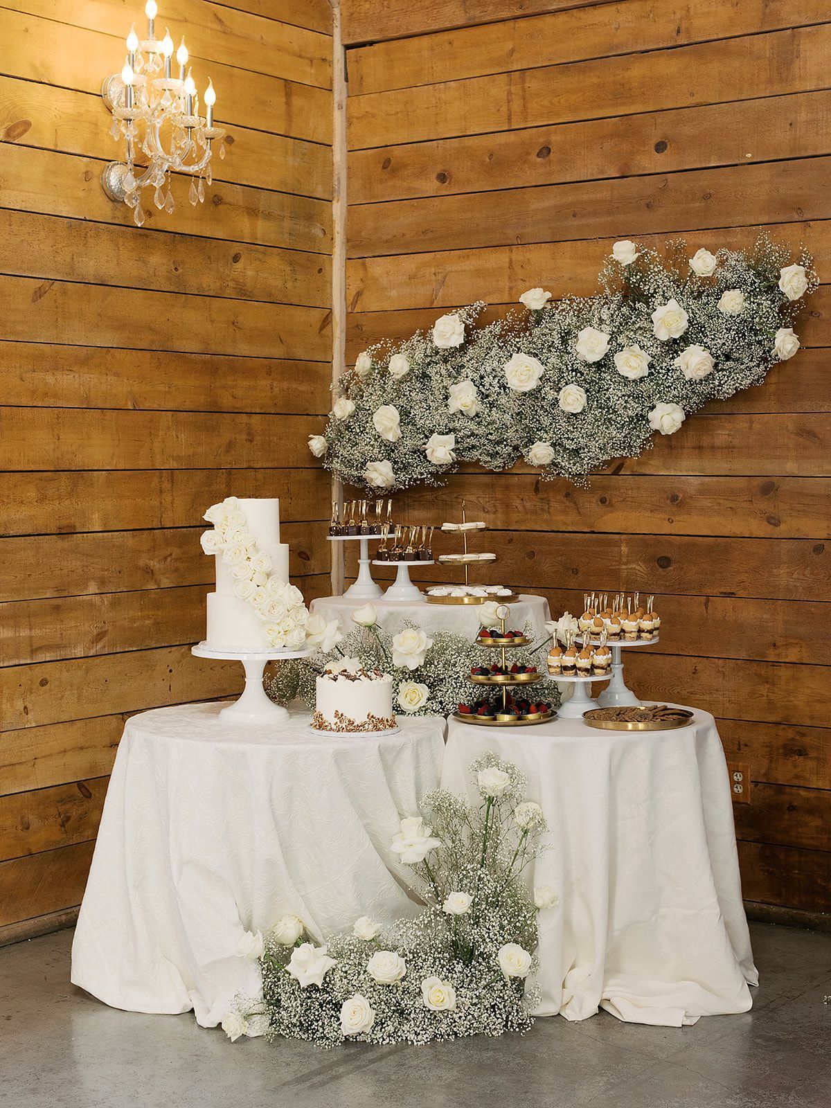 White dessert table with cakes and flowers at a rustic wooden wall wedding reception.