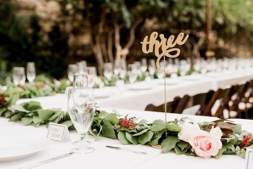 Elegant outdoor wedding reception table with white linens, green garlands, and a gold “three” table sign