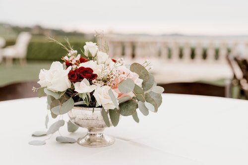 White and red floral centerpiece in a glass vase on a table, with a blurred bridge in the background