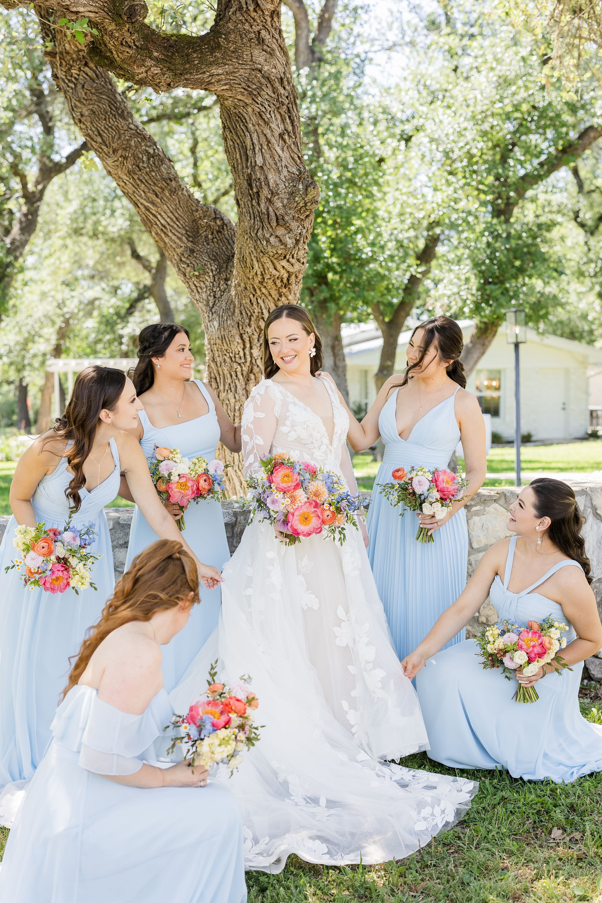 Bride with bridesmaids in light blue dresses holding colorful bouquets outdoors under a tree