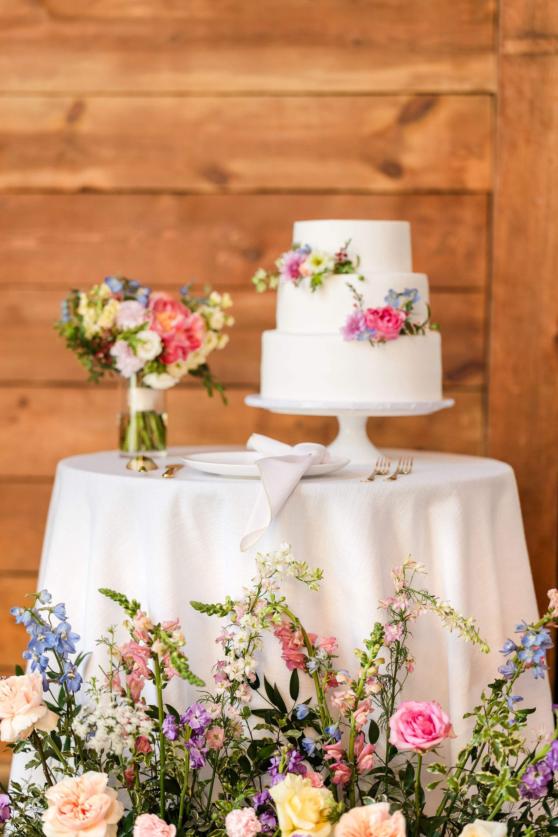Two-tier white wedding cake with pink flowers on a draped table, framed by pastel floral arrangements.