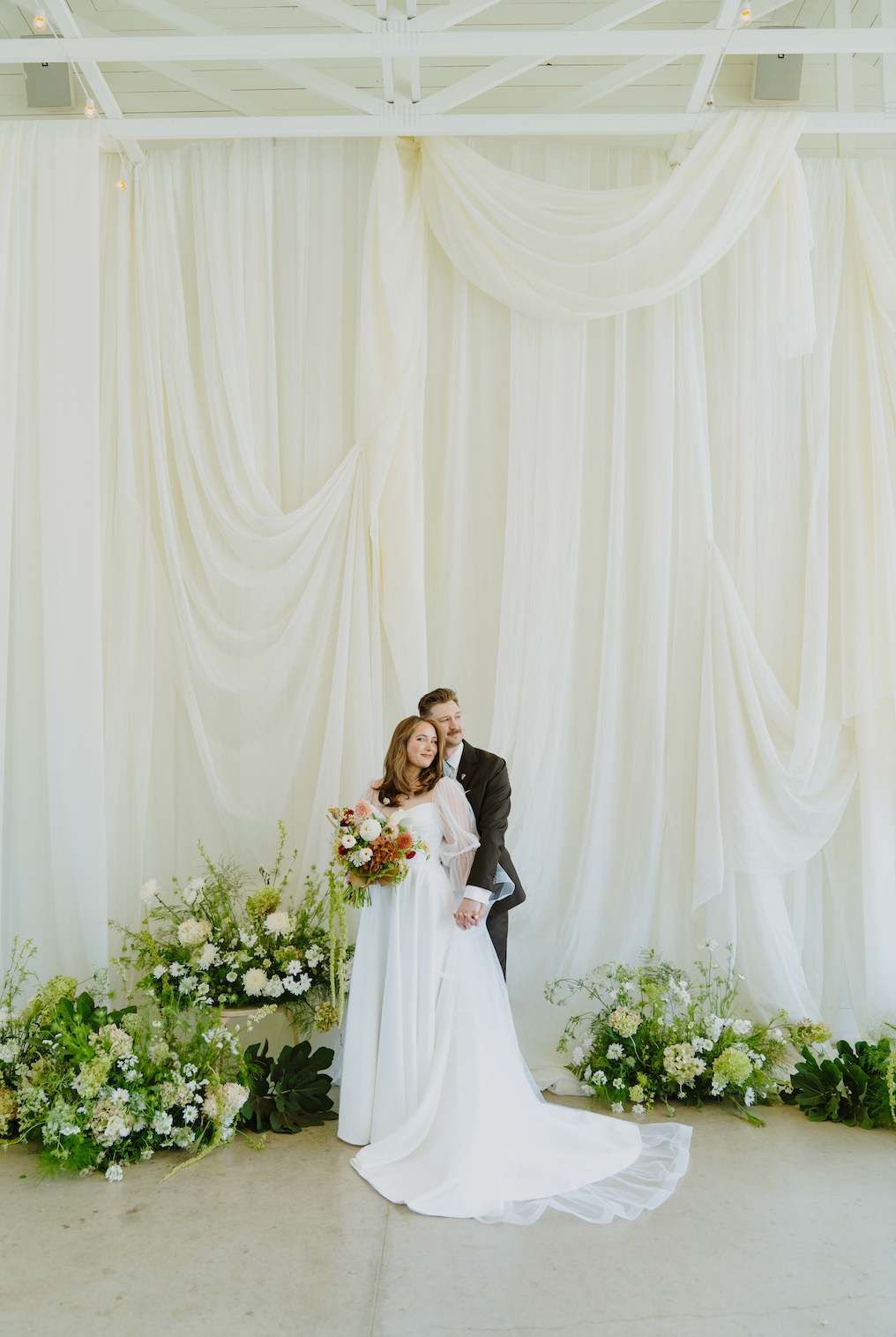 Bride and groom embracing before a white draped backdrop with floral arrangements