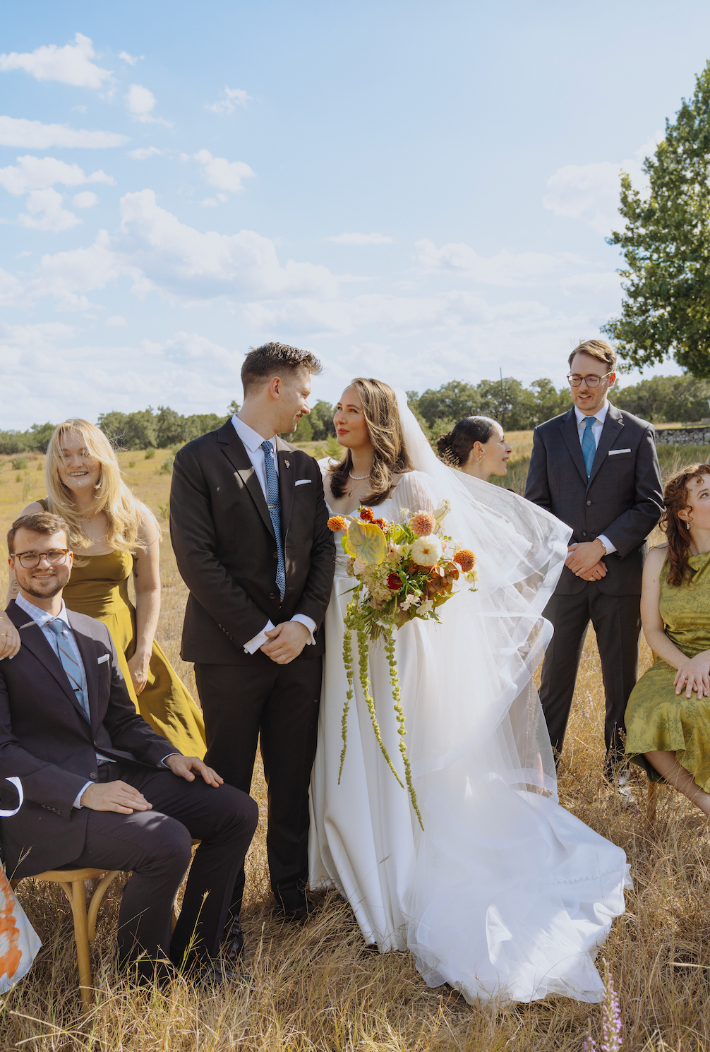 Wedding party outdoors in a field, bride in white holding bouquet beside groom in a black suit