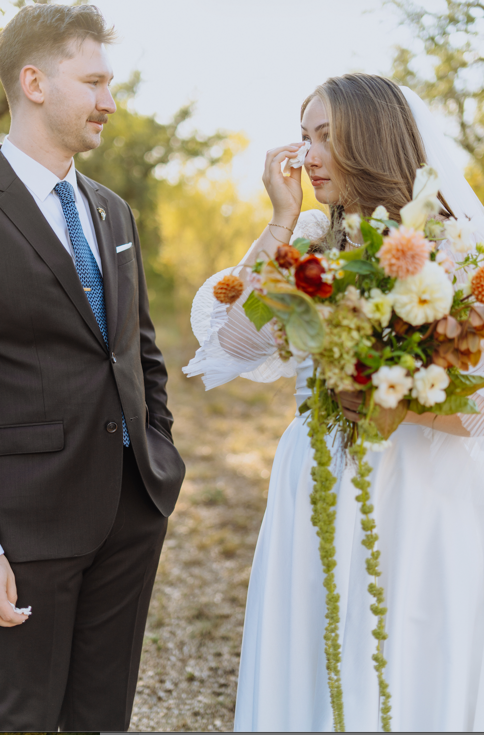 Bride holding a bouquet adjusts her hair while standing beside a groom outdoors in a sunlit field.