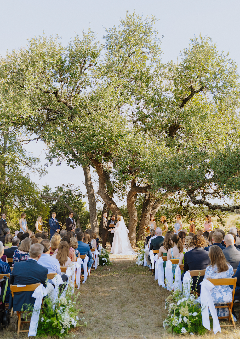 Outdoor wedding ceremony under large trees with guests seated along a grassy aisle and a bride in white at center