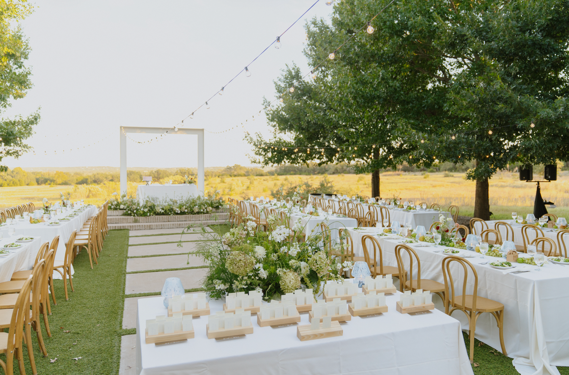Outdoor wedding reception setup with white tables, gold chairs, floral centerpieces, and a tree-lined field backdrop