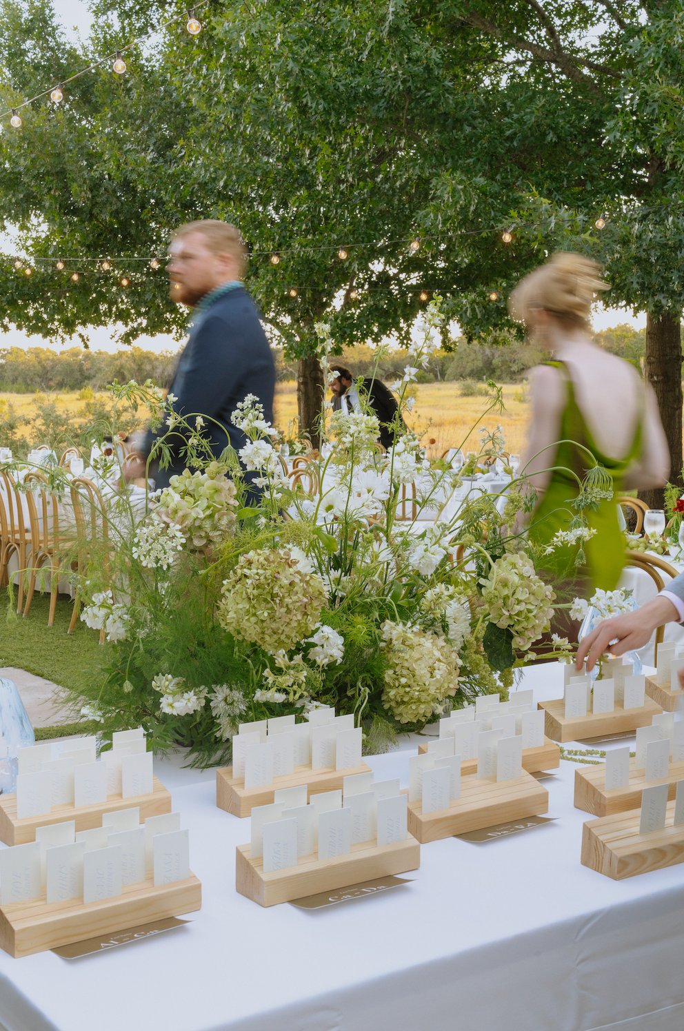 Outdoor reception table with white and gold decor, greenery centerpiece, and blurred guests walking behind