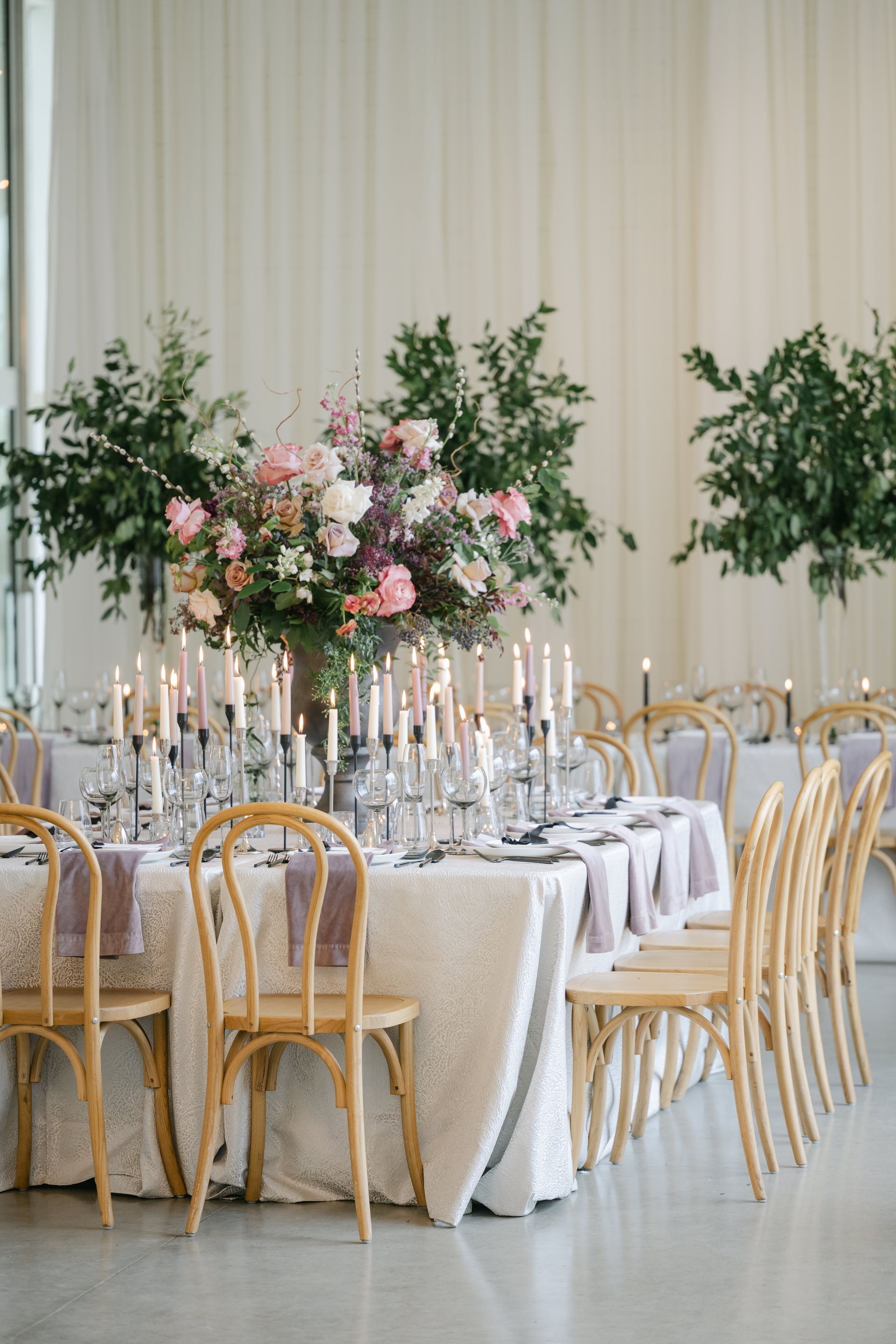 Elegant wedding reception table with white chairs, candles, and pink floral centerpieces in a bright hall