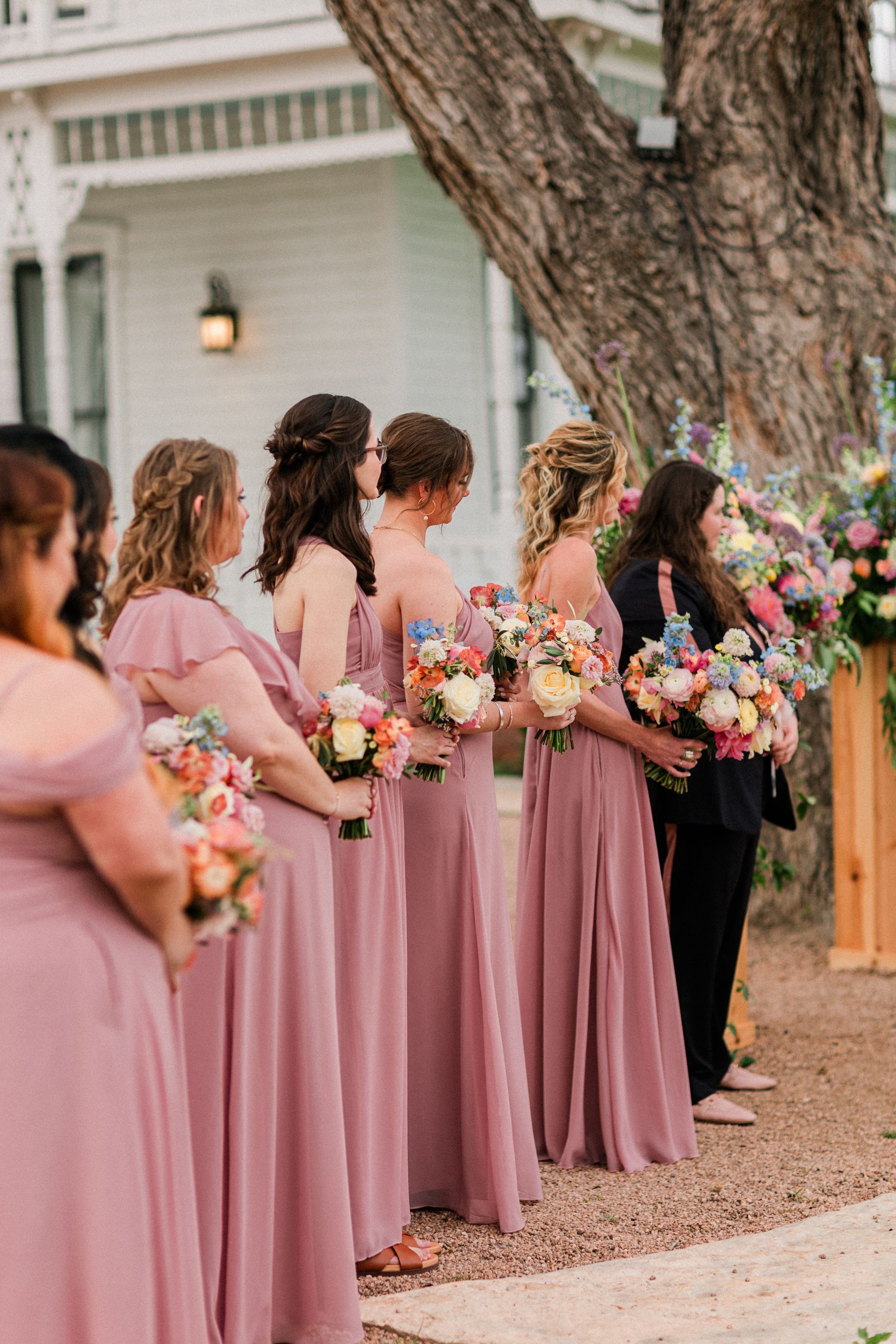 Bridesmaids in blush dresses holding flower bouquets stand outdoors beside a tree and house.