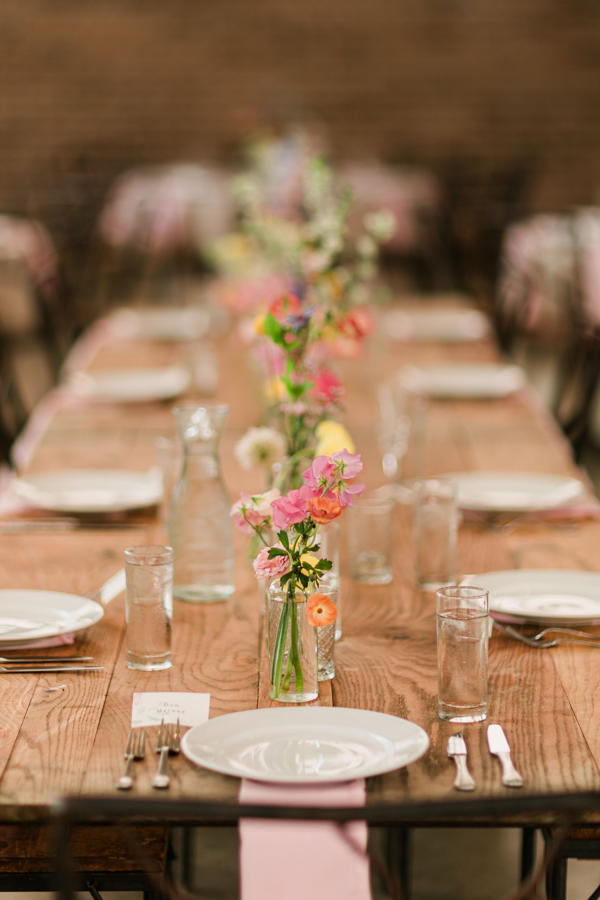 Long wooden dining table set with plates, glasses, and pink floral centerpieces