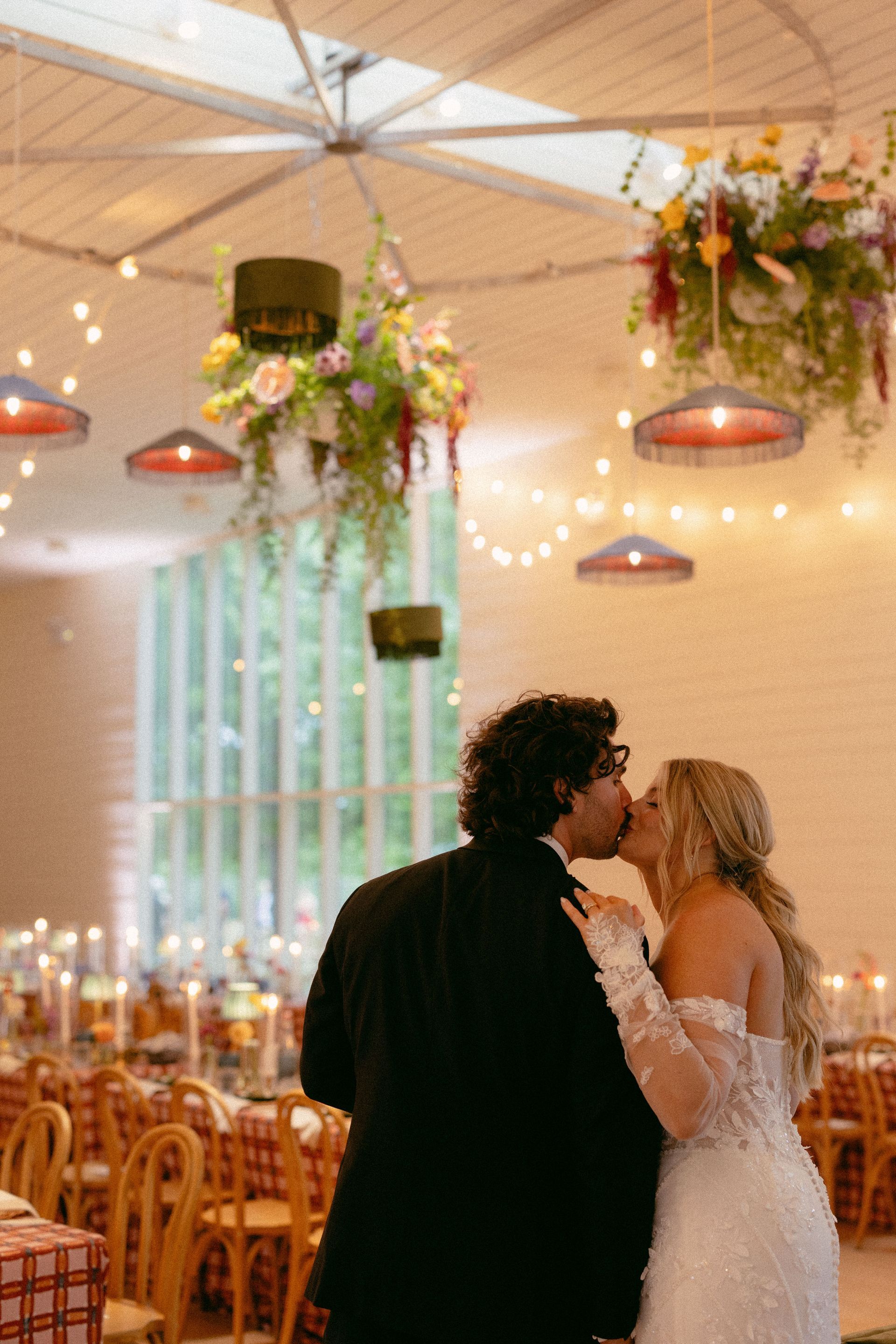 Newlyweds kiss at a decorated indoor reception with hanging flowers and warm pendant lights