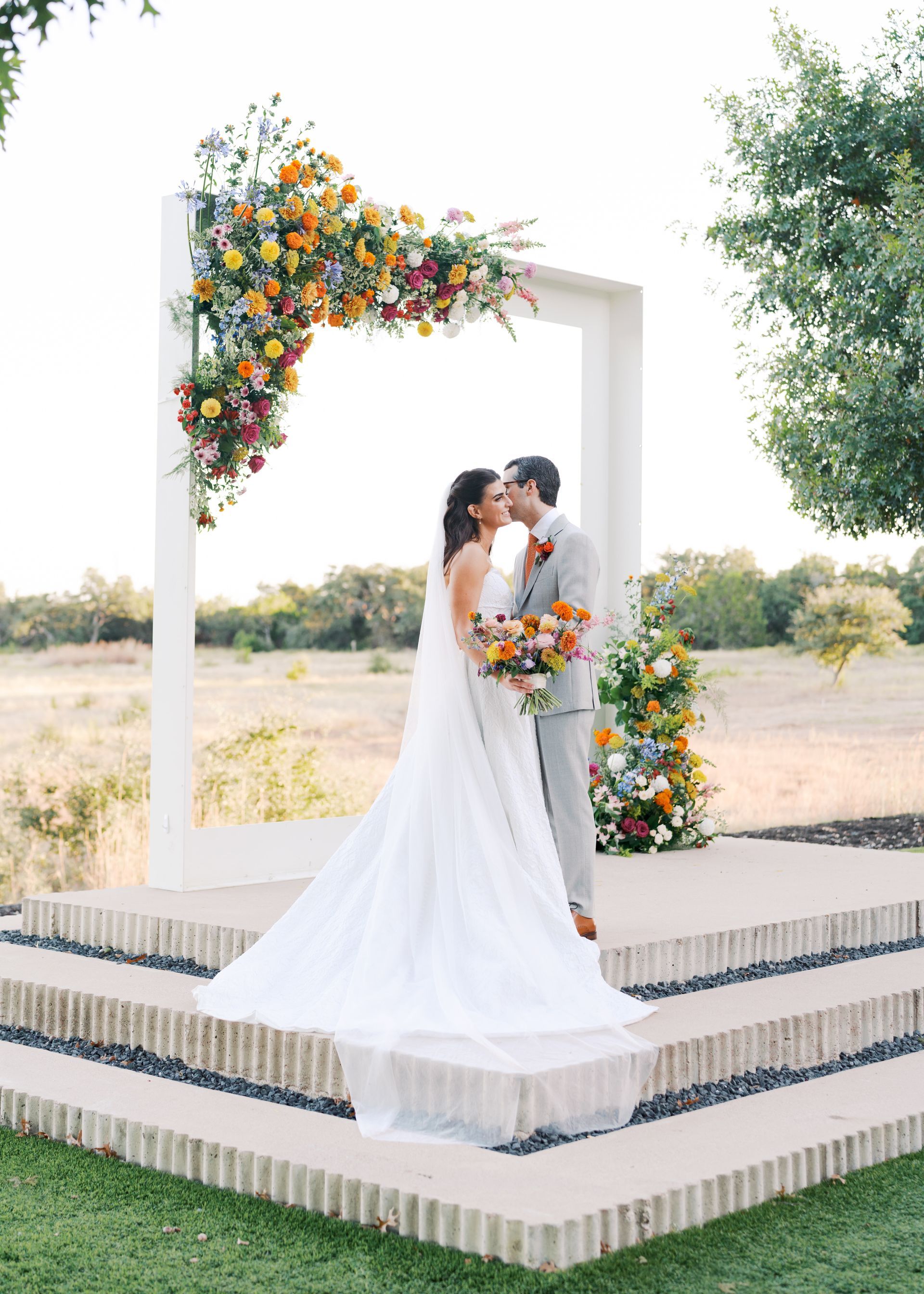 Couple kissing on a floral-decorated wedding platform outdoors at sunset