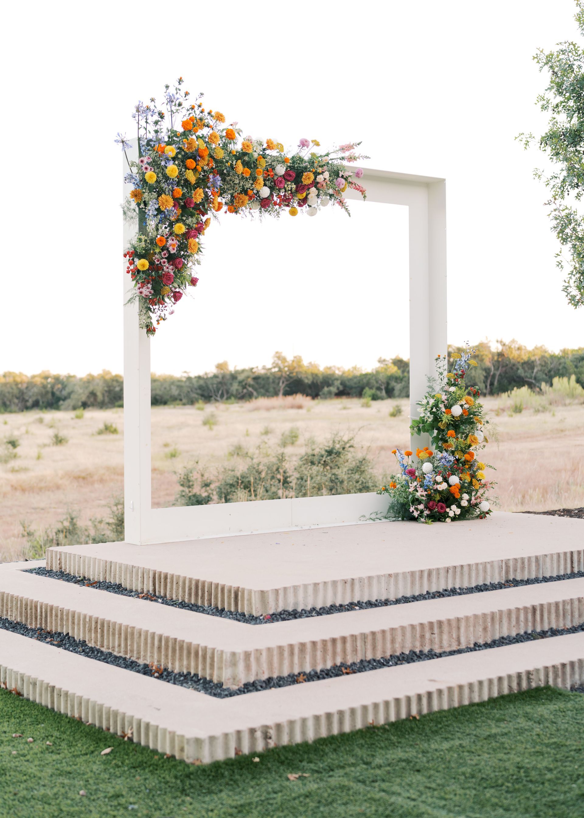 Floral-decorated white wedding stage outdoors with layered platform and grassy foreground