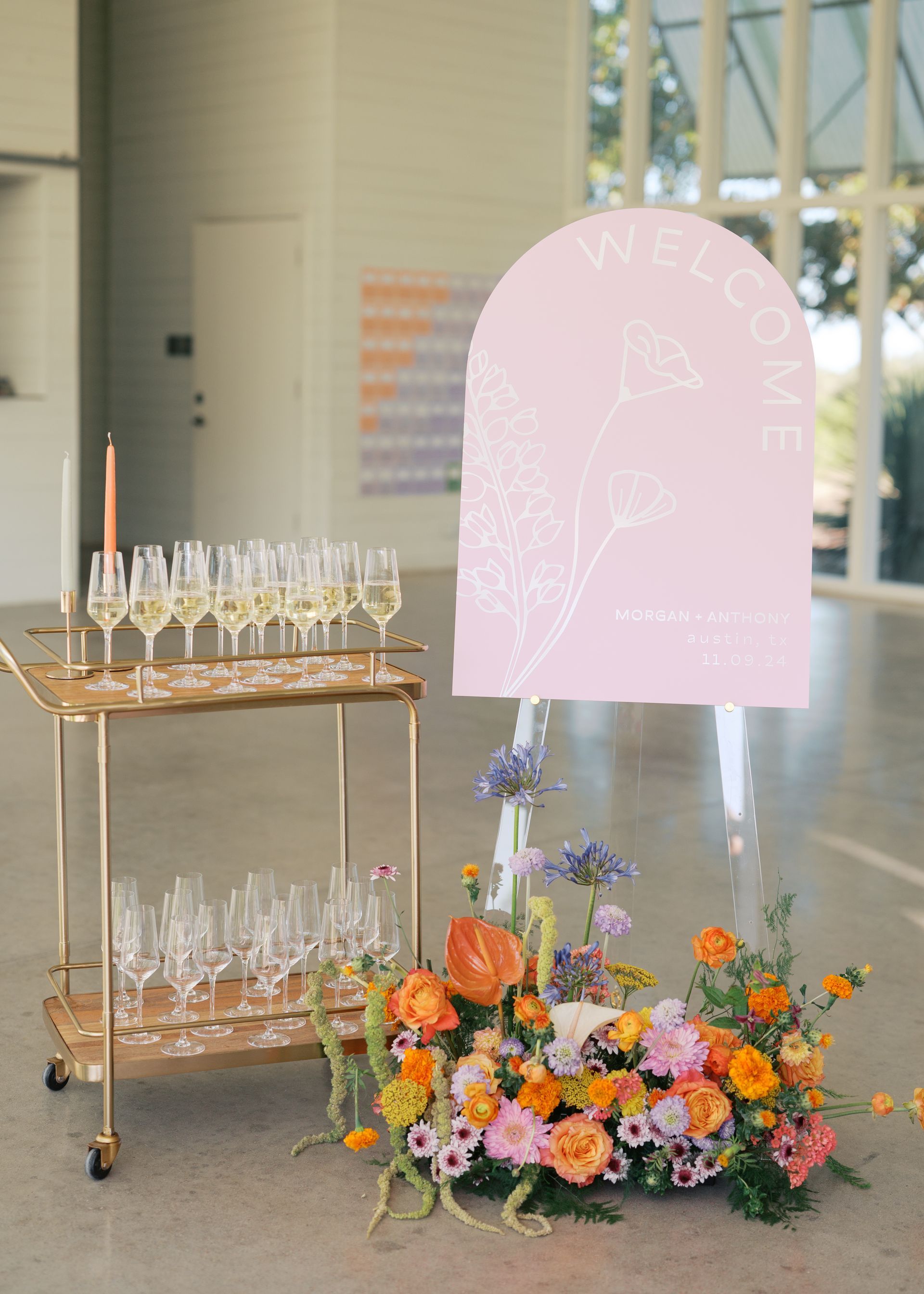 Floral event display with champagne glasses on a gold cart beside a pastel pink sign and flower arrangement.