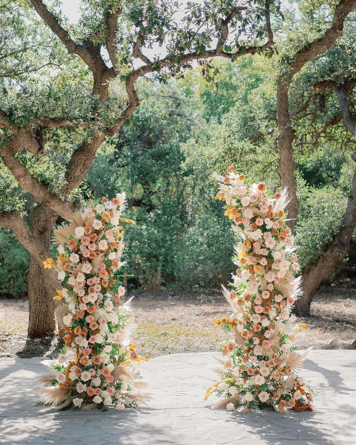 Two floral pillars of peach and white flowers in a garden path under trees