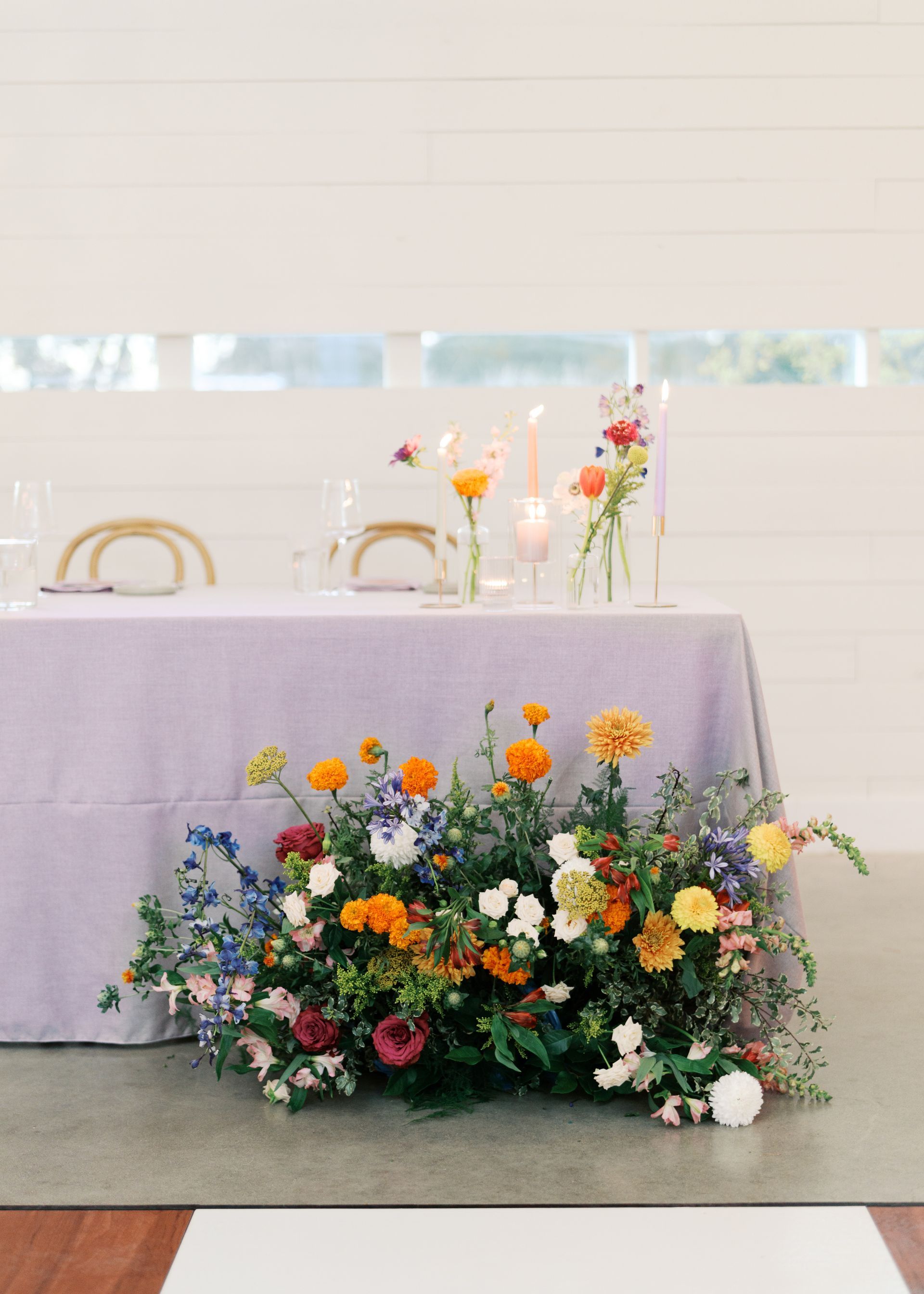 Flower arrangements on a table and floor in a bright, minimalist room with white chairs and a lavender tablecloth