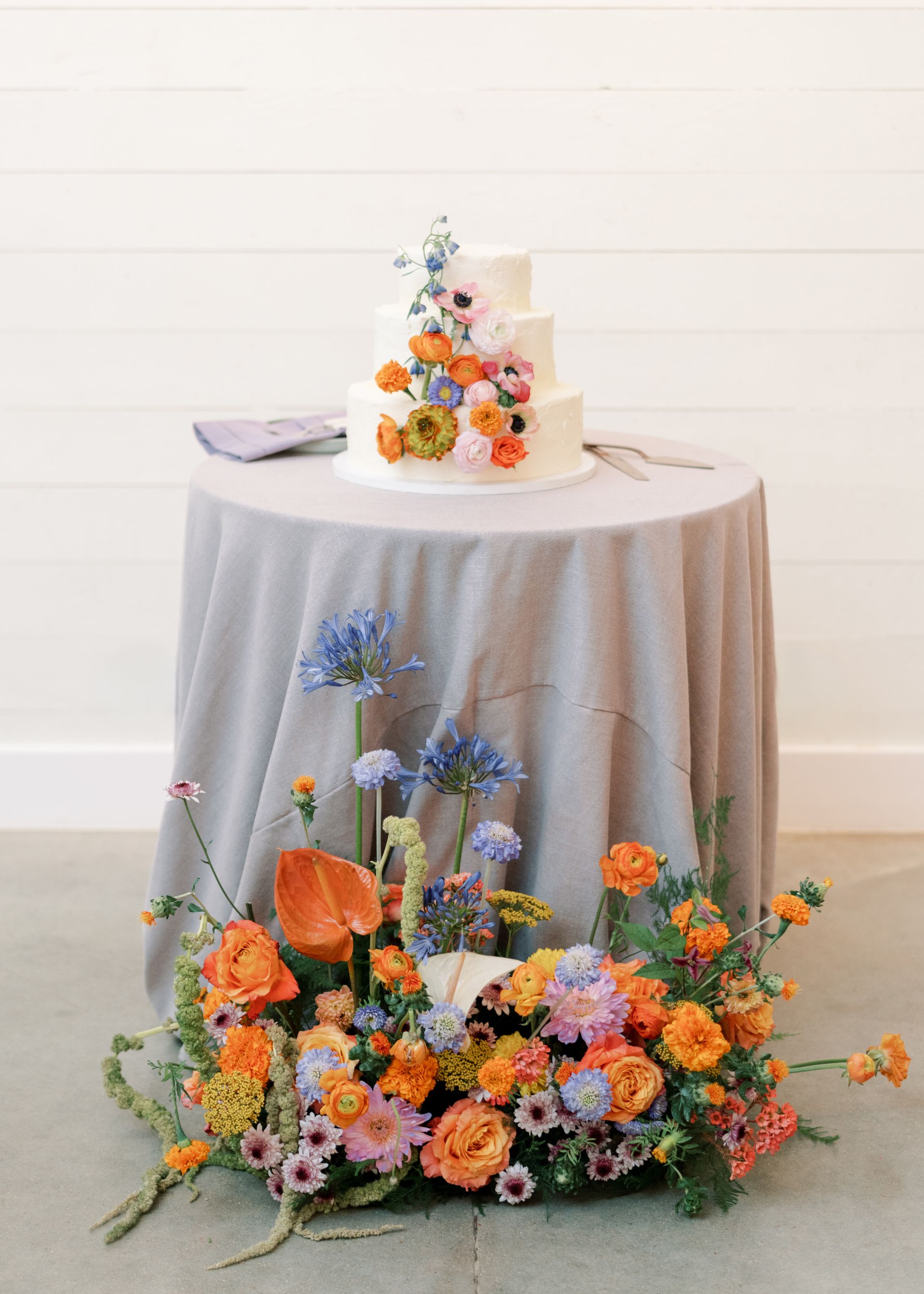 Flower-adorned cake on a round table with a white cloth, surrounded by colorful floral arrangements.