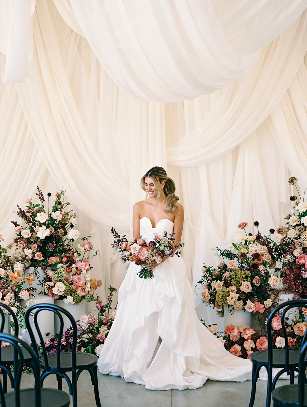 Bride in a white gown holding a bouquet at a floral wedding backdrop with draped ivory curtains
