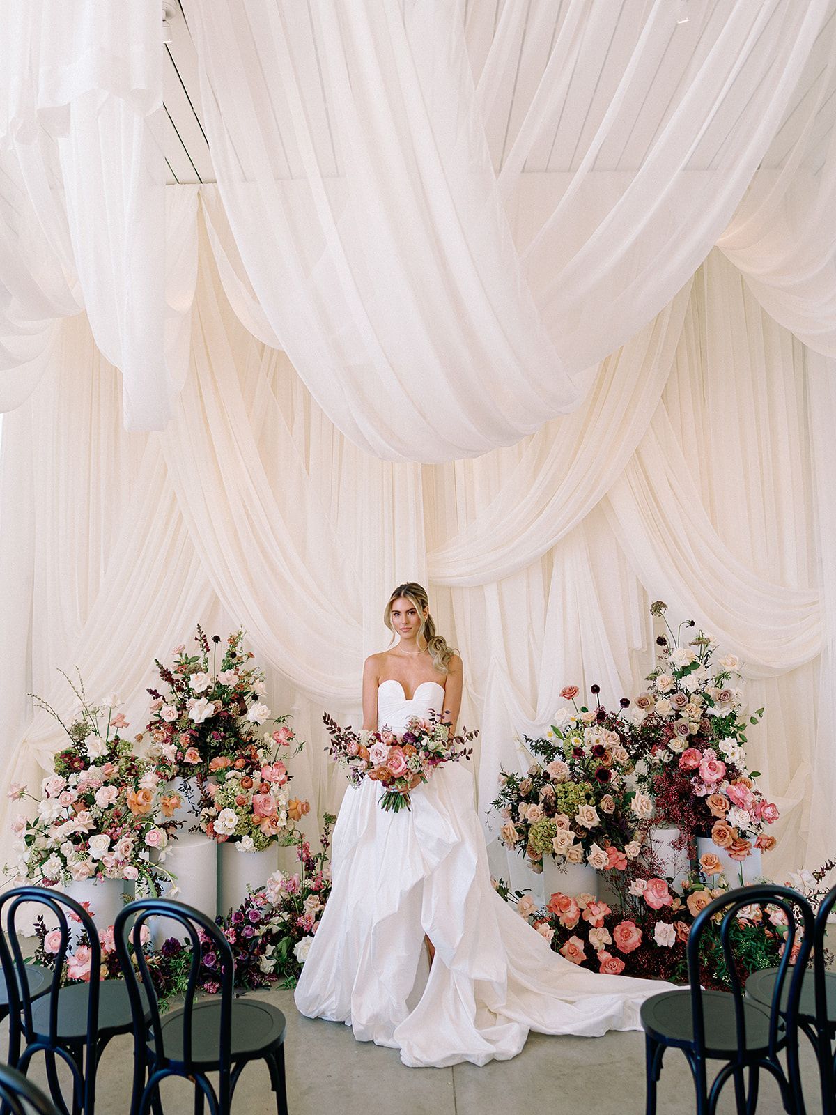 Woman in a white gown standing between floral arrangements at a wedding backdrop