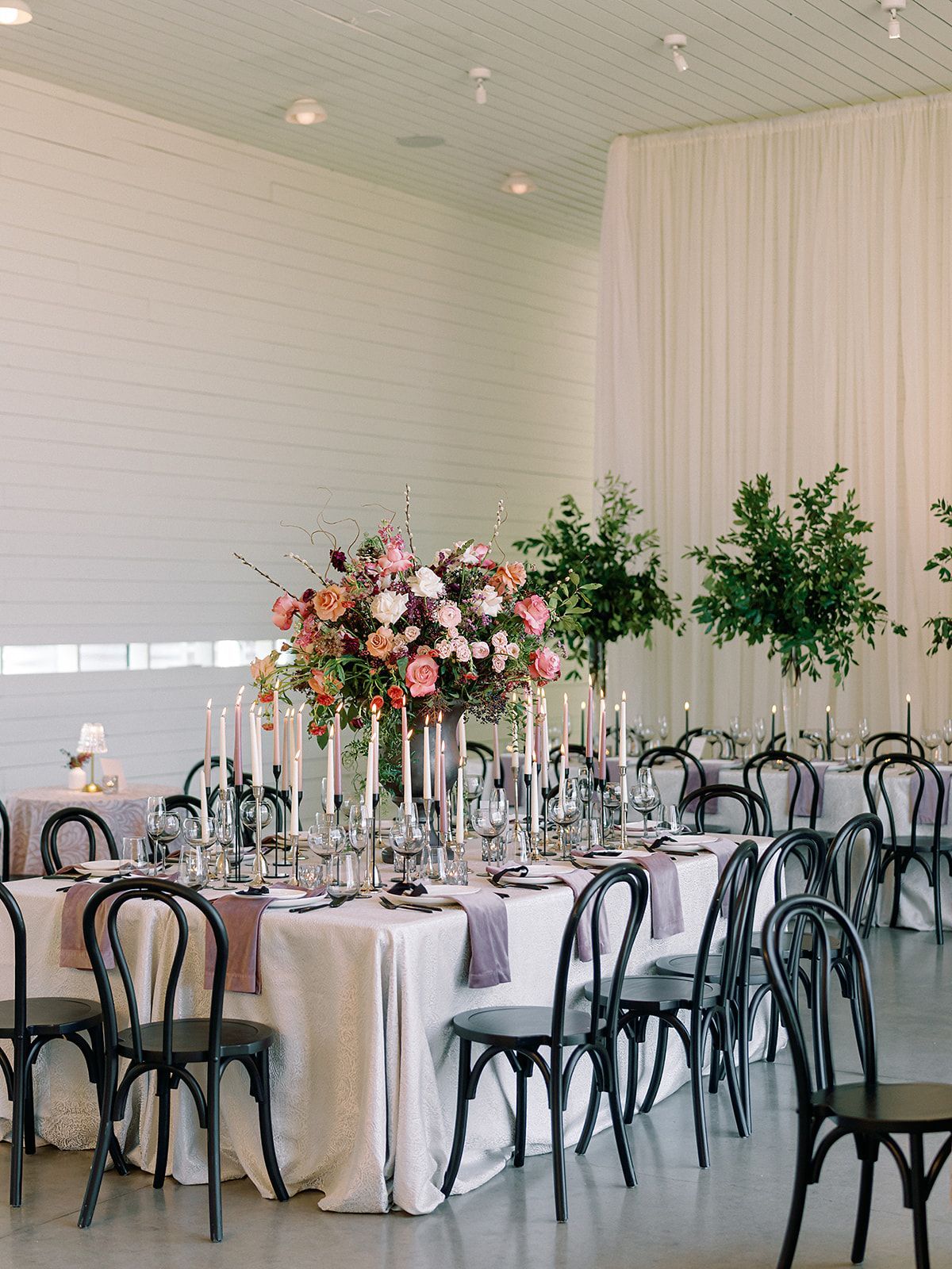 Elegant banquet table with white linens, pink floral centerpiece, and black chairs in a bright hall