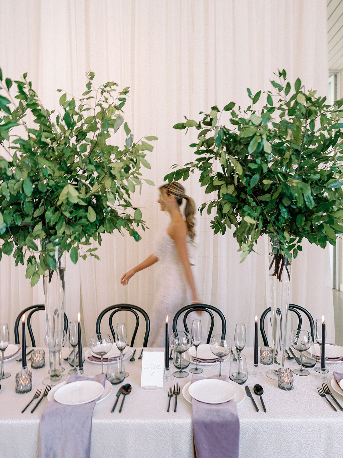 Elegant banquet table with black chairs, green floral arrangements, and a person in a tan dress in the background