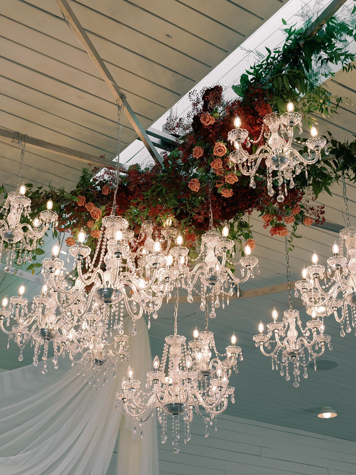Elegant crystal chandeliers hang beneath a draped ceiling with green and burgundy floral garlands.