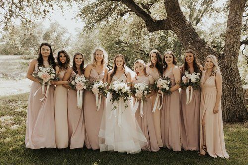 Bridesmaids in blush dresses and a bride in white holding bouquets under a tree outdoors