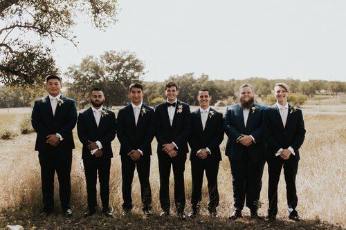 Seven men in black suits standing in a grassy field outdoors