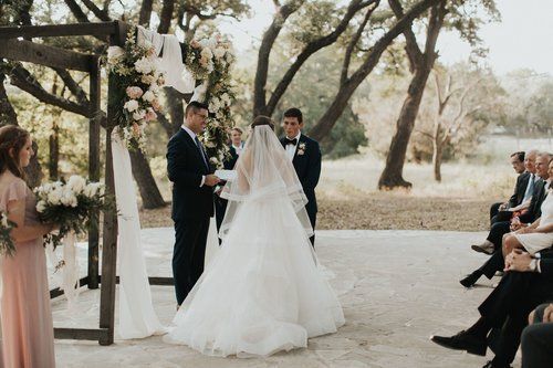 Wedding ceremony outdoors beneath trees, with bride in white gown and groom exchanging vows before seated guests.