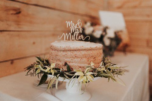 Rustic wedding cake with floral greenery and “Mr & Mrs Miller” topper on a white table
