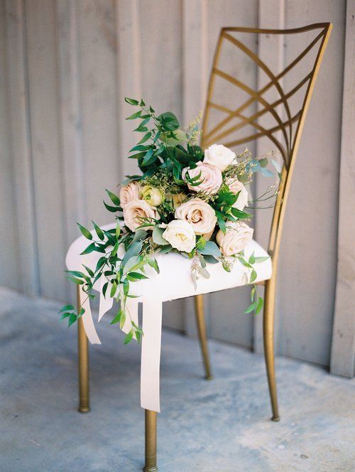 White chair with gold legs decorated with white and peach flowers and greenery against a light backdrop