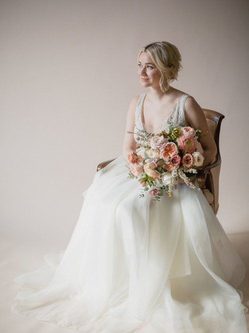 Bride in a white gown holding a bouquet of blush and peach flowers while seated by a plain wall