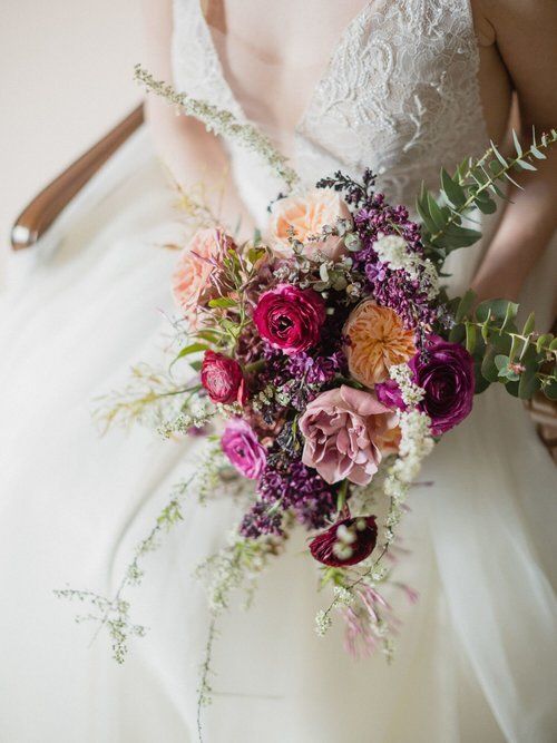 Bride holding a colorful bouquet of pink, purple, and peach flowers in a white lace wedding dress