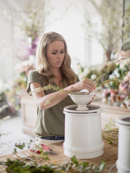 Woman arranging flowers in a bright studio, placing blooms into a white pedestal vase