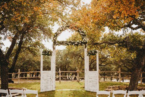 Outdoor wedding arch under autumn trees in a grassy park, with white chairs in the foreground