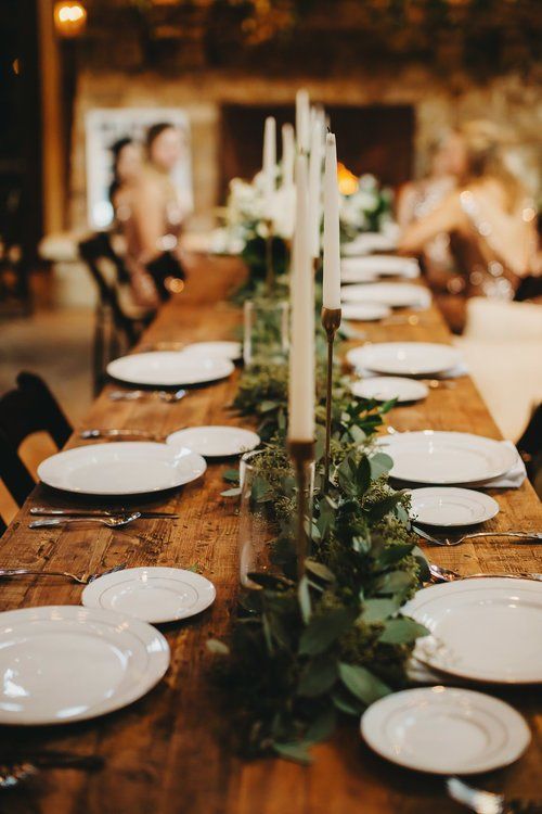 Elegant banquet table with white plates, candles, and greenery centerpiece in a warmly lit room