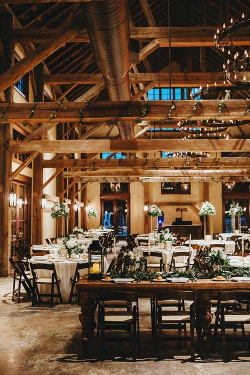 Rustic banquet hall with long wooden tables, white chairs, floral centerpieces, and warm string lights