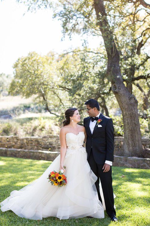 Bride and groom standing together outdoors beneath a tree, with the bride holding a colorful bouquet.