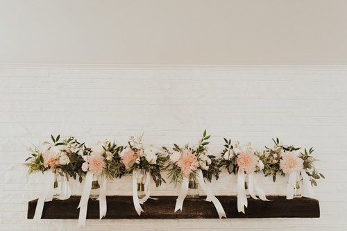 White floral arrangement with pink roses and hanging ribbons on a dark wooden shelf against a white wall